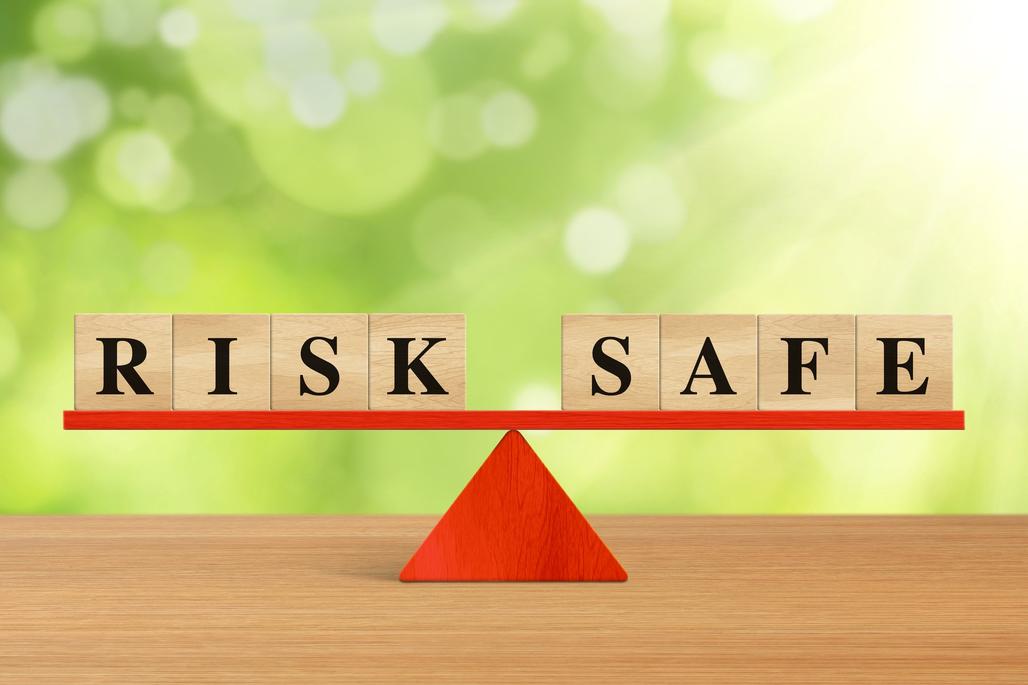 The words risk and safe spelled out in wooden block letters being balanced on a red seesaw sitting on a wooden table.