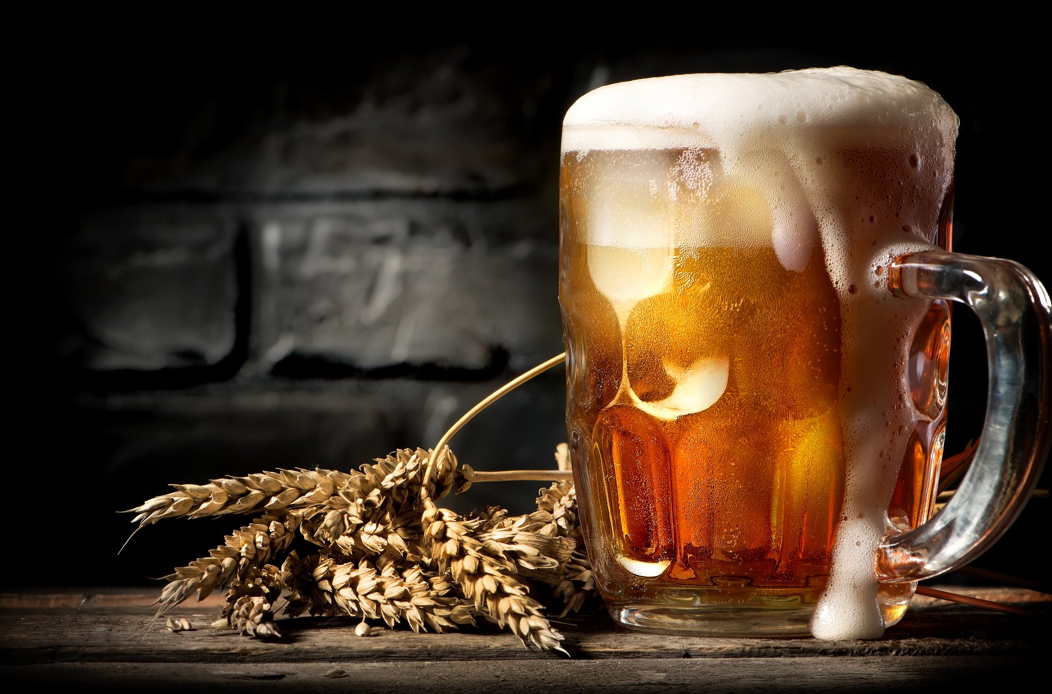 A large, overflowing glass of beer with a stem of wheat lying on a table.