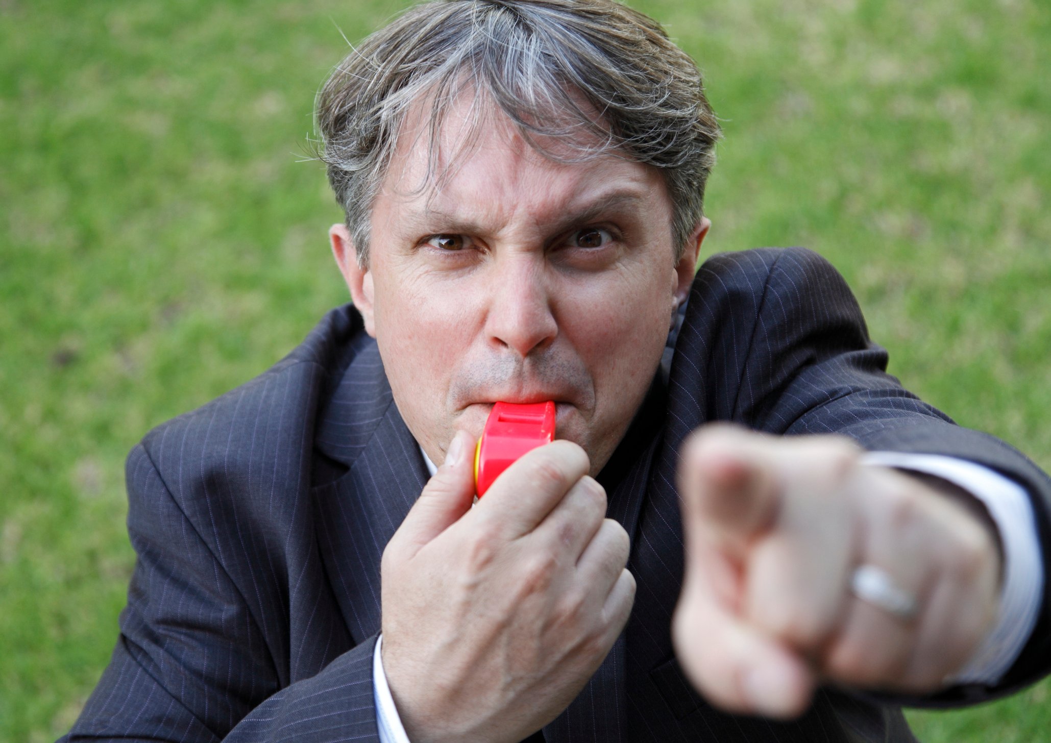 A man in a suit with an angry look pointing his finger and blowing a whistle.