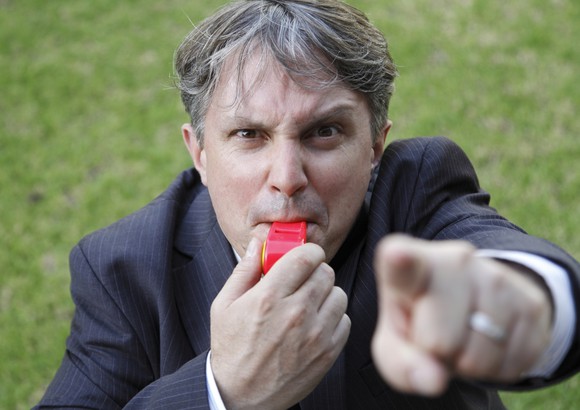 A man in a suit with an angry look pointing his finger and blowing a whistle.
