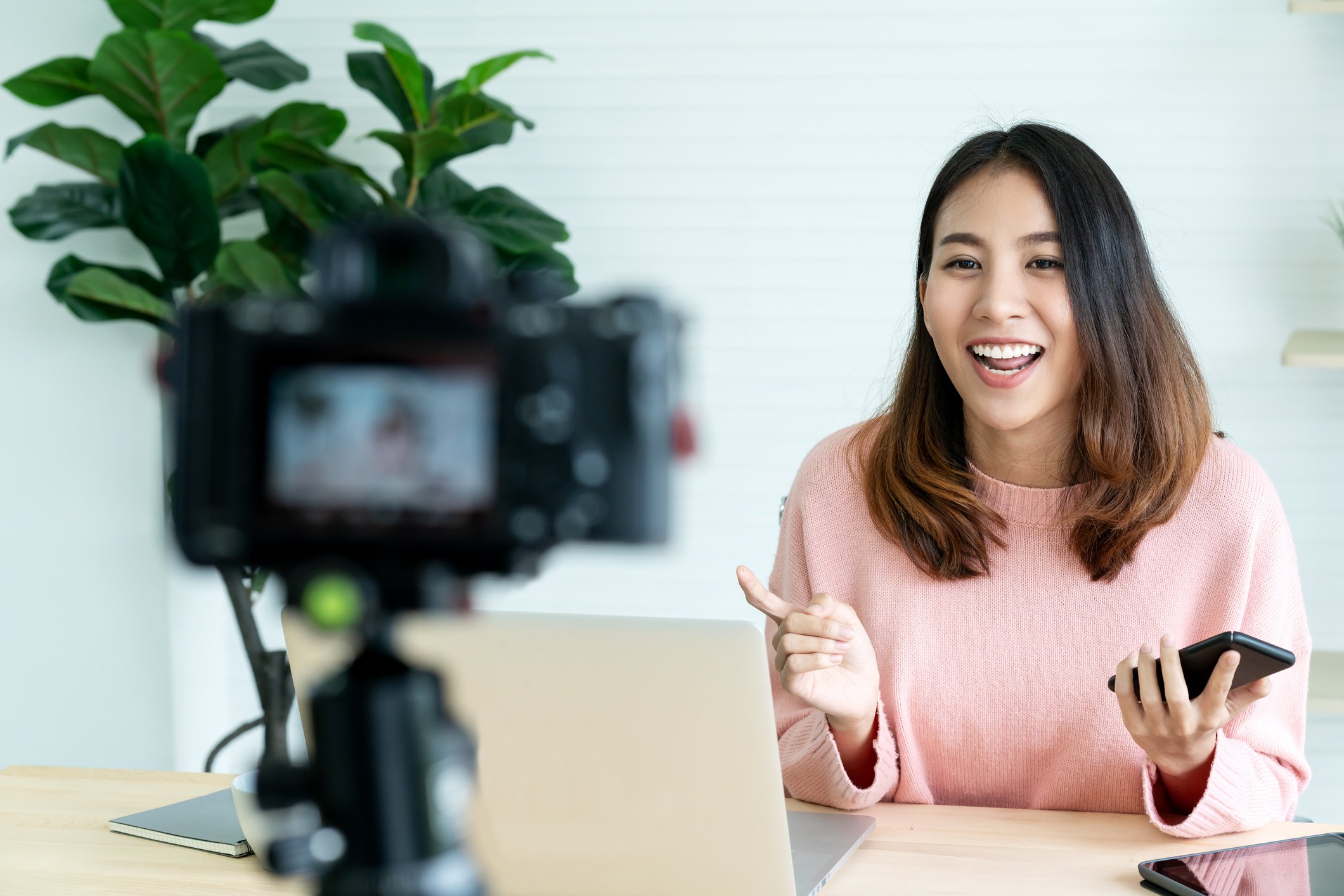 Young asian woman smiles and speaks into a mounted camera taking a video of her behind a desk.