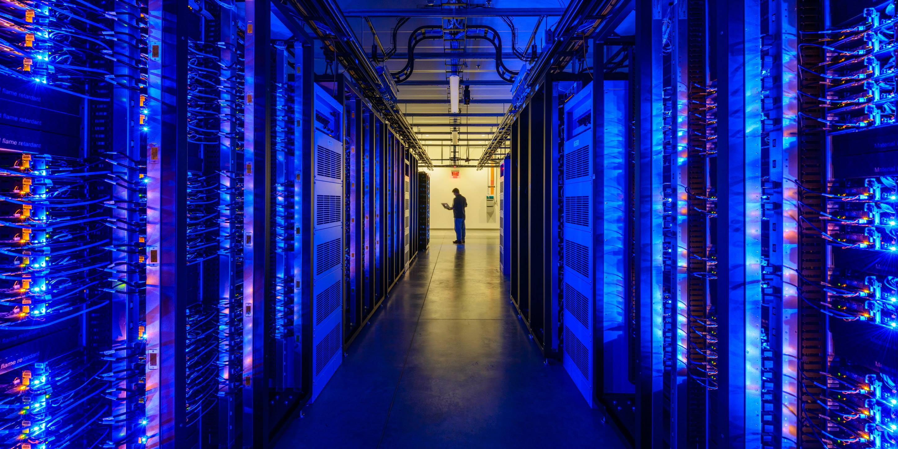 A darkened hall filled with server racks on either side and a silhouette of a Facebook worker at the end, in the company's Prineville, Oregon, data center.