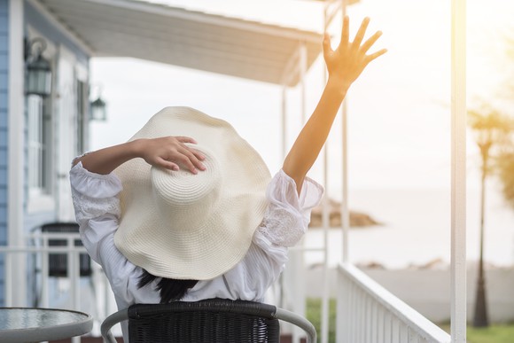 A seated woman wearing a broad-brimmed hat, with her left hand on the top of her hat and her right hand up in the air.