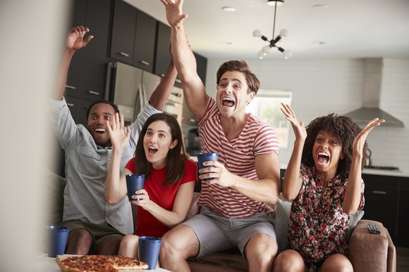 A group of twenty-something friends cheer and react to a game on TV as they drink from cups and a pizza is in front of them on a table. 