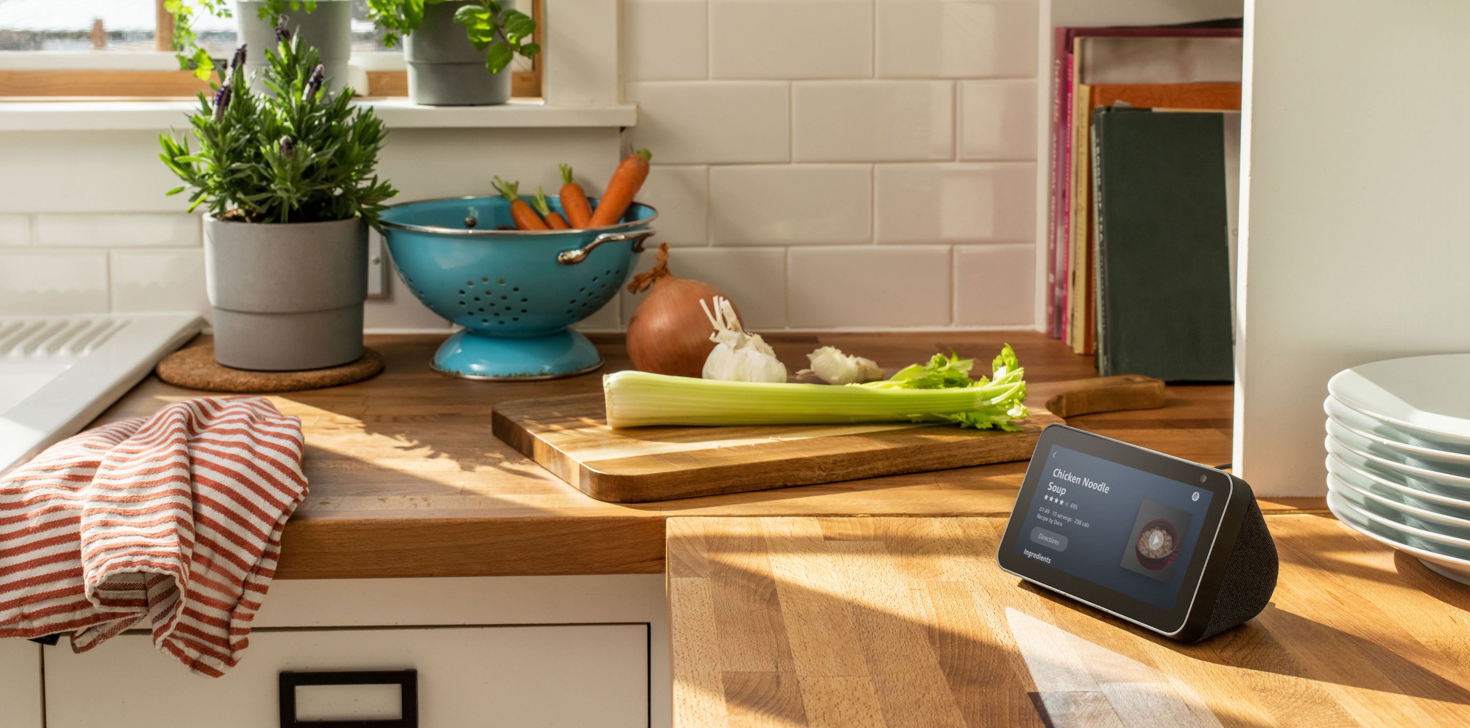 Echo Show 5 on a kitchen counter next to a cutting board with vegetables on it and a colander containing carrots