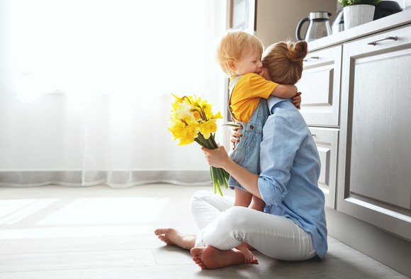 Woman sitting on floor in a kitchen, hugging young child while holding flowers.