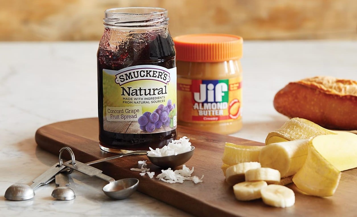 Jars of jelly and peanut butter next to banana and bread.