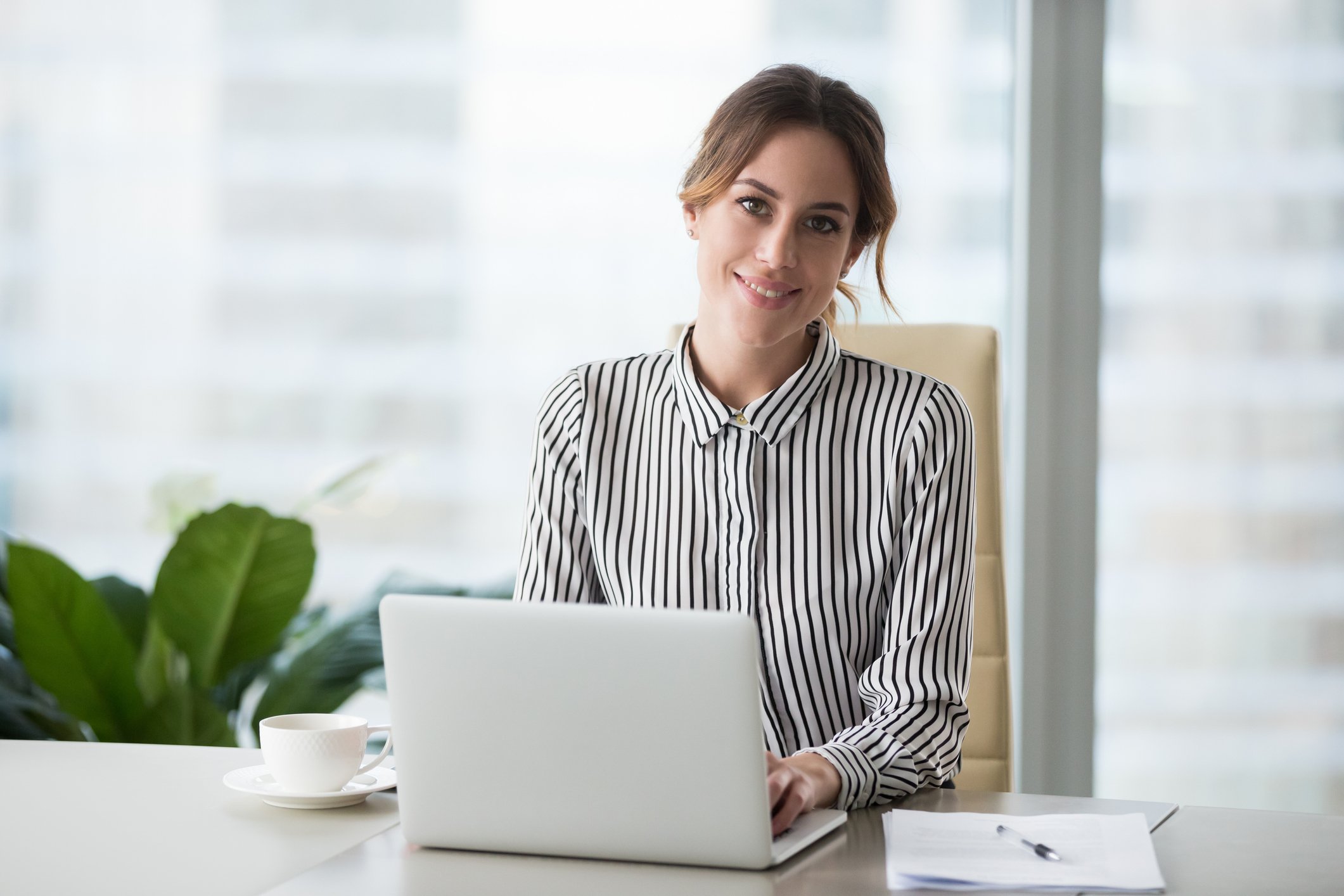A woman in business casual attire sits at a desk, with an open laptop in front of her and a picture window behind her.