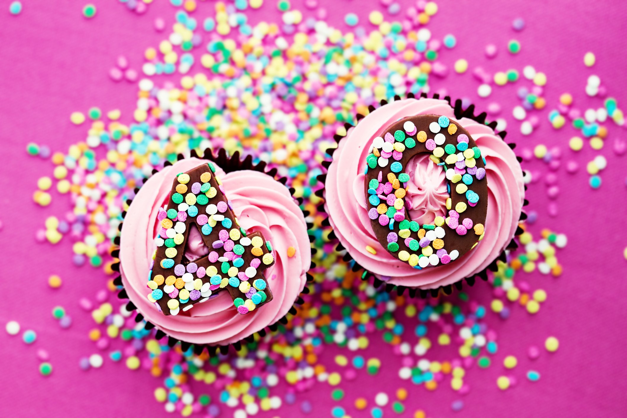 Two cupcakes, on a pink surface adorned with colorful sprinkles, with the number 40 written in sprinkles on top of them.