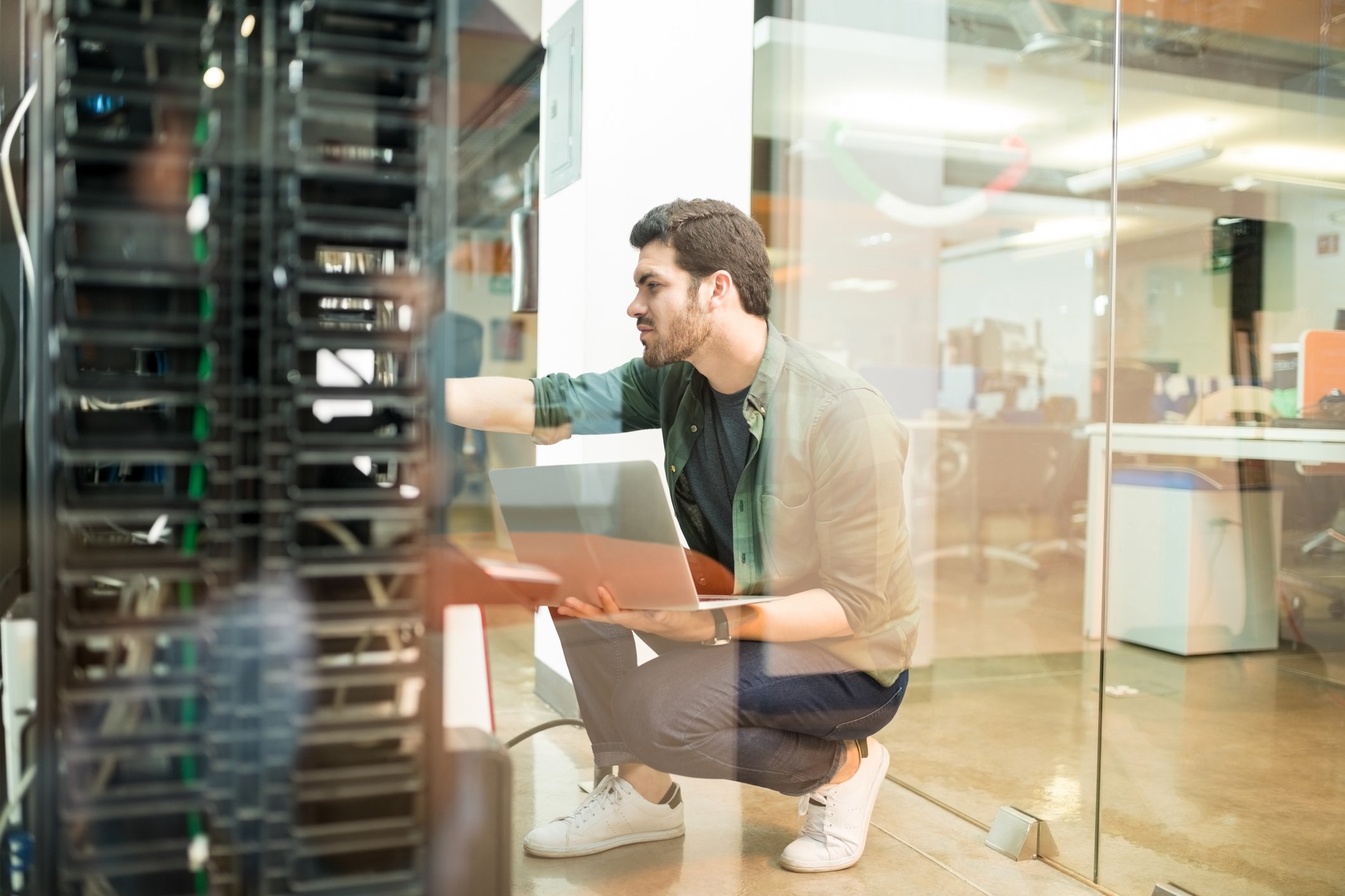 A young man holds a laptop while kneeling and checking out a rack of servers in an IT room.