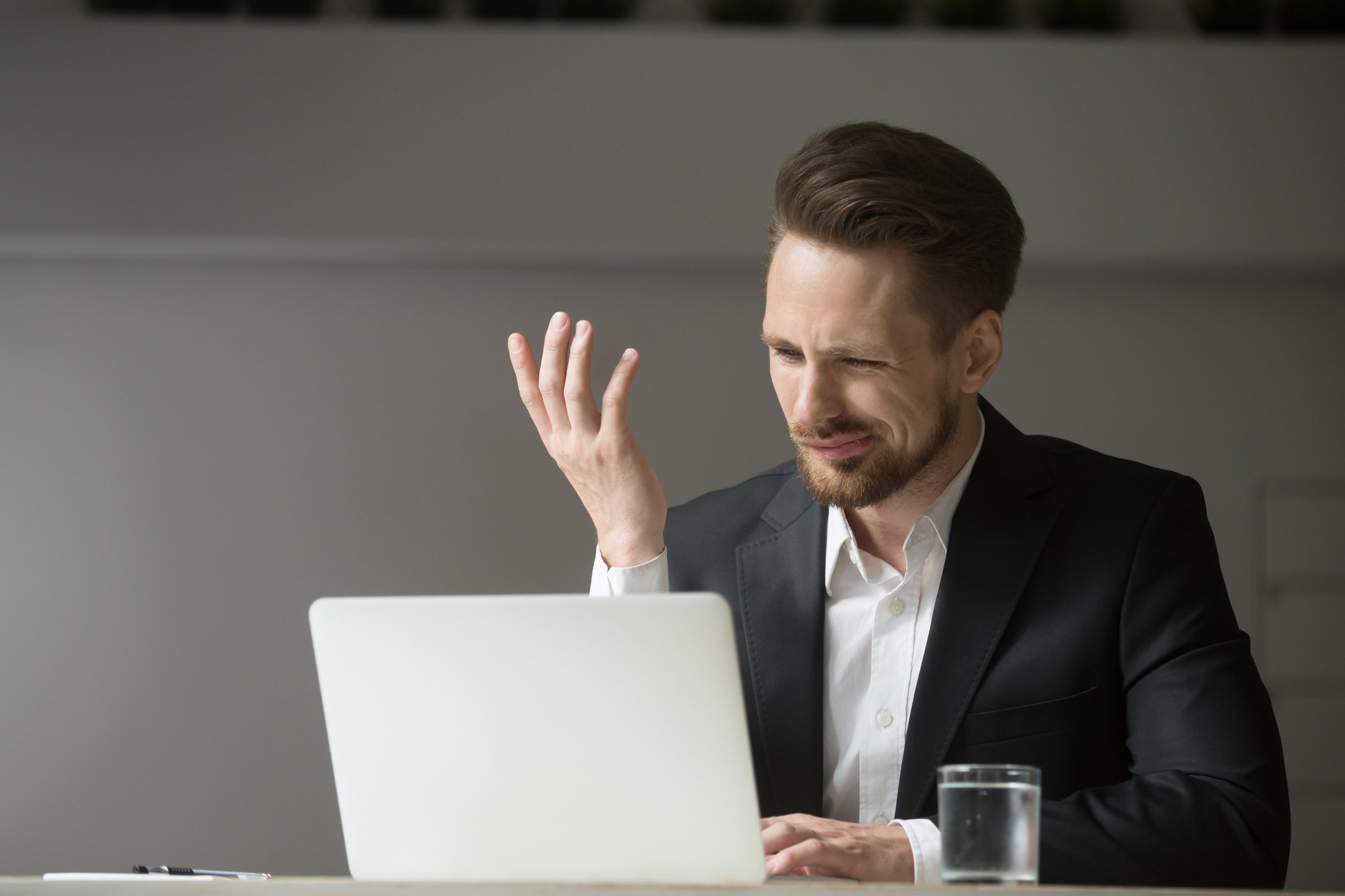 A young man with a beard in a suit winces at something on his laptop. 