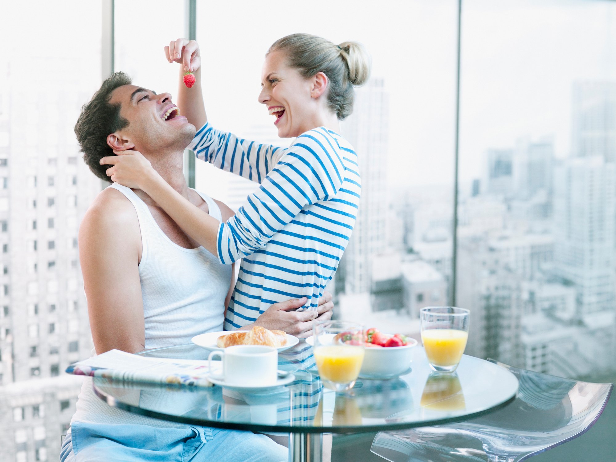 A couple sitting at a breakfast table in their apartment. The woman is feeding the man a strawberry.