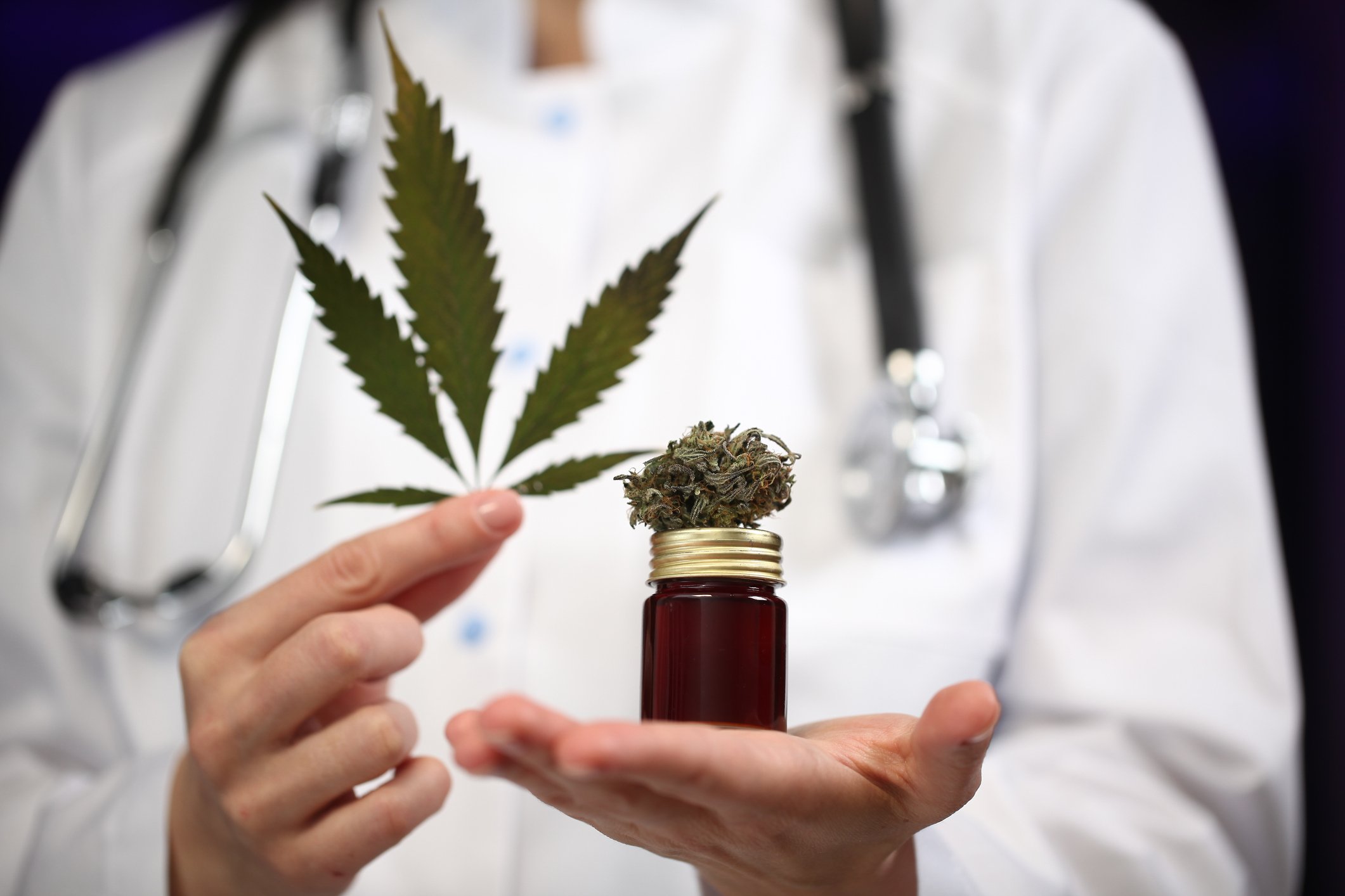 Person in lab coat and stethoscope holds a marijuana leaf and small jar of cannabis.