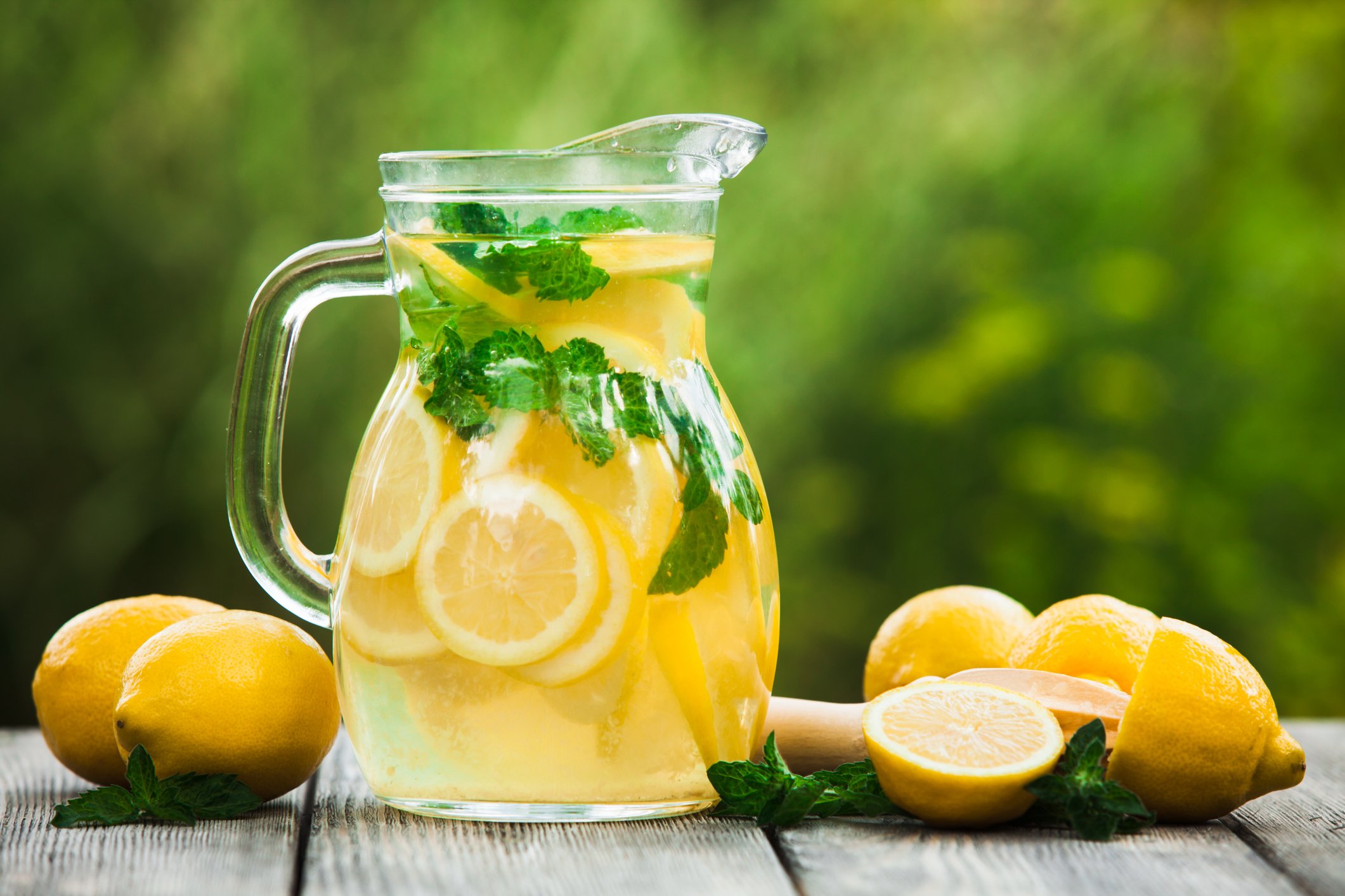 A pitcher of lemonade surrounded by fresh lemons on a wood table outdoors.
