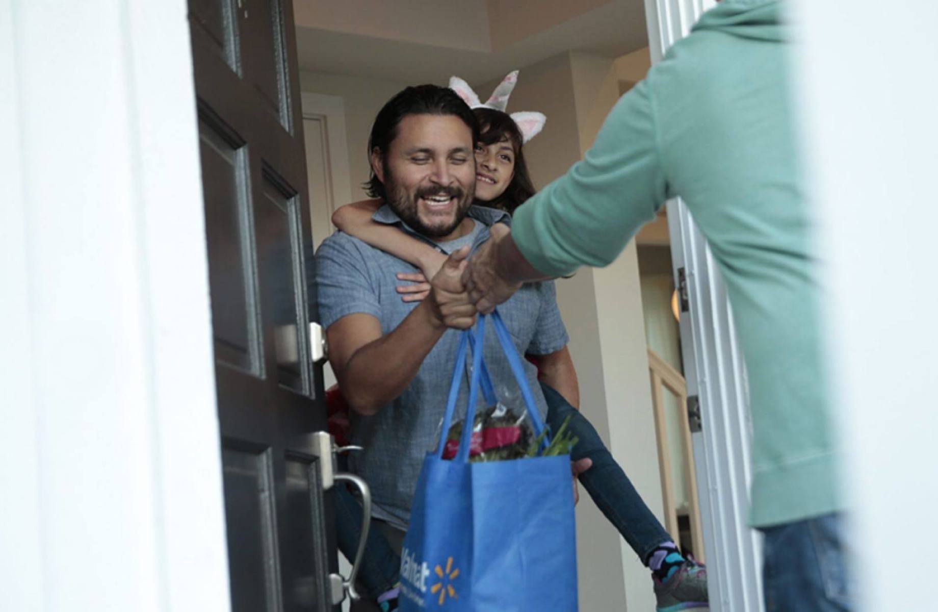 A man with a child on his back receiving a grocery bag from someone at the front door.