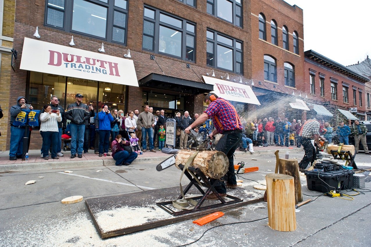 Person using chainsaw to cut log on a street in front of a Duluth Trading Company store.