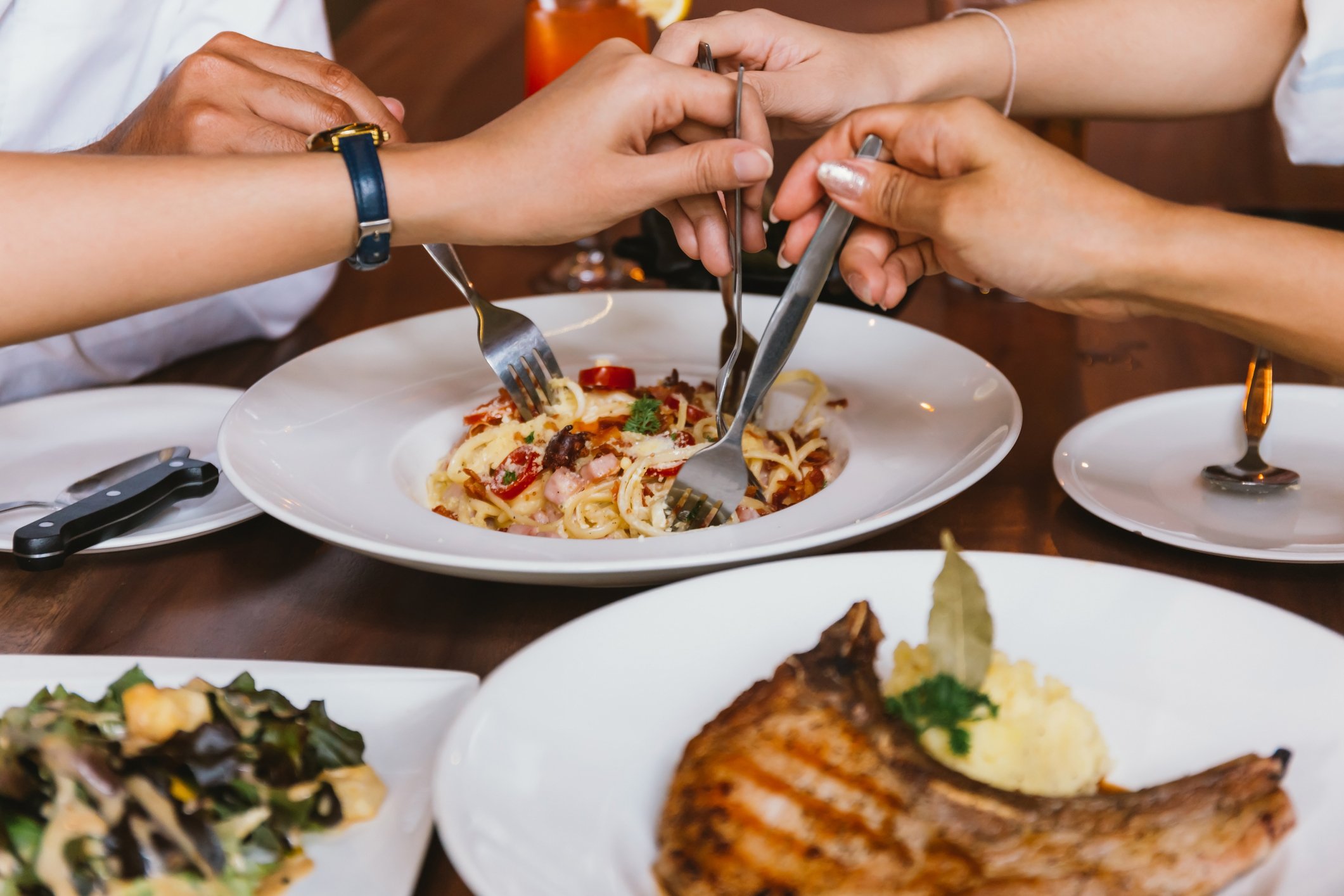 Friends plunge their forks into a shared plate of pasta at a fast-casual restaurant.
