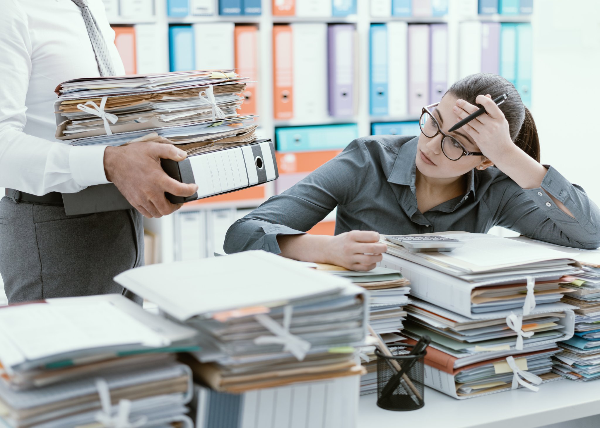 A person sits at a table piled with papers looking exasperated.