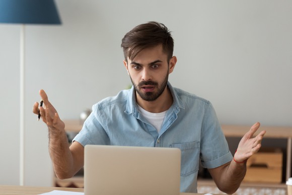 A man shrugs at his laptop with a befuddled expression on his bearded face.