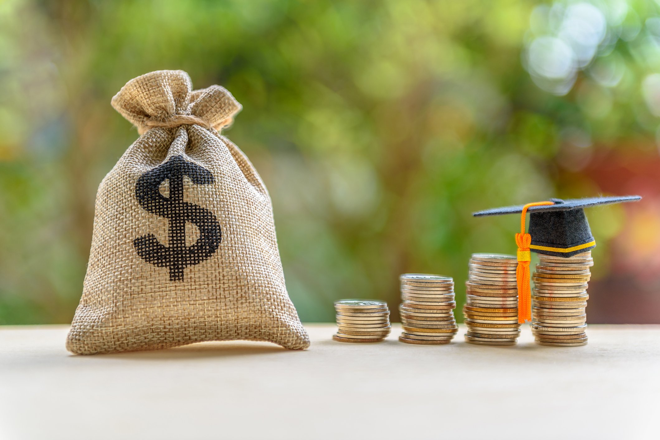 Bag of money sitting next to coins, atop of which is a graduation cap.