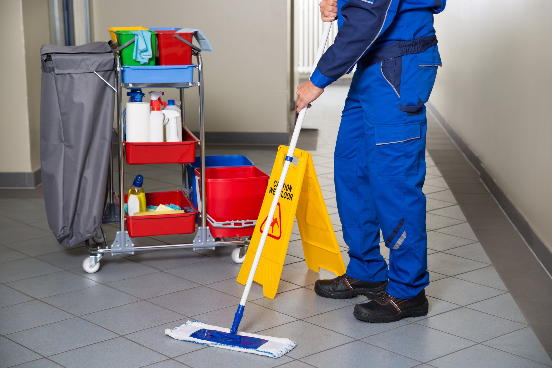 A custodian in a blue uniform mops an office corridor with his supplies cart nearby.