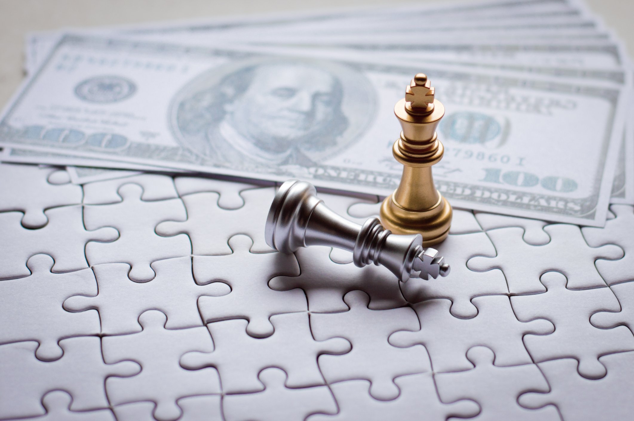 Gold chess king and fallen silver chess king next to hundred dollar bills, all on top of a jigsaw puzzle