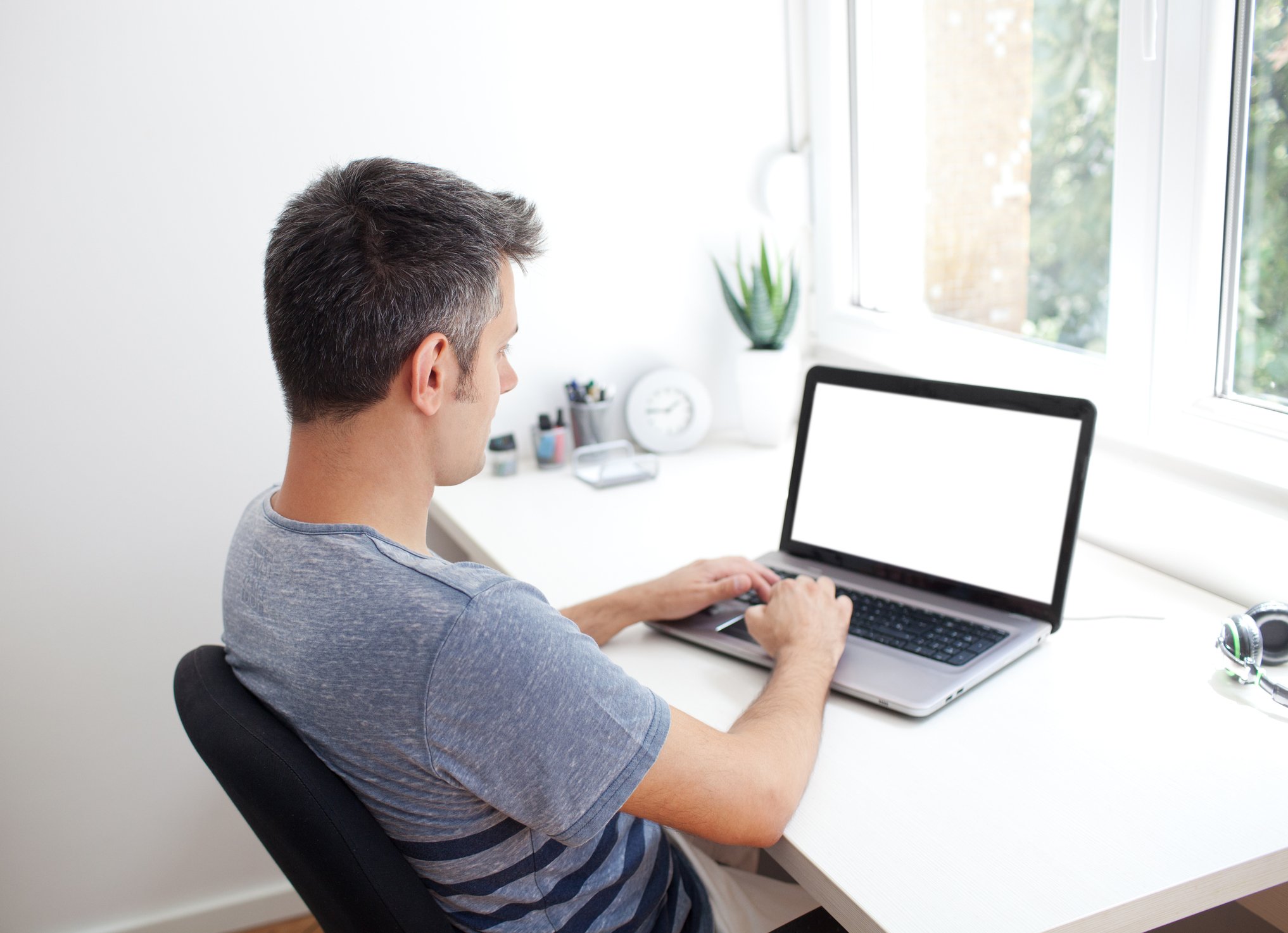 A man in a T-shirt works on a laptop, which is on a table near a window.