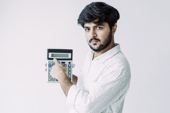 An unhappy young man with a beard points to a calculator as if showing the numbers to you. 