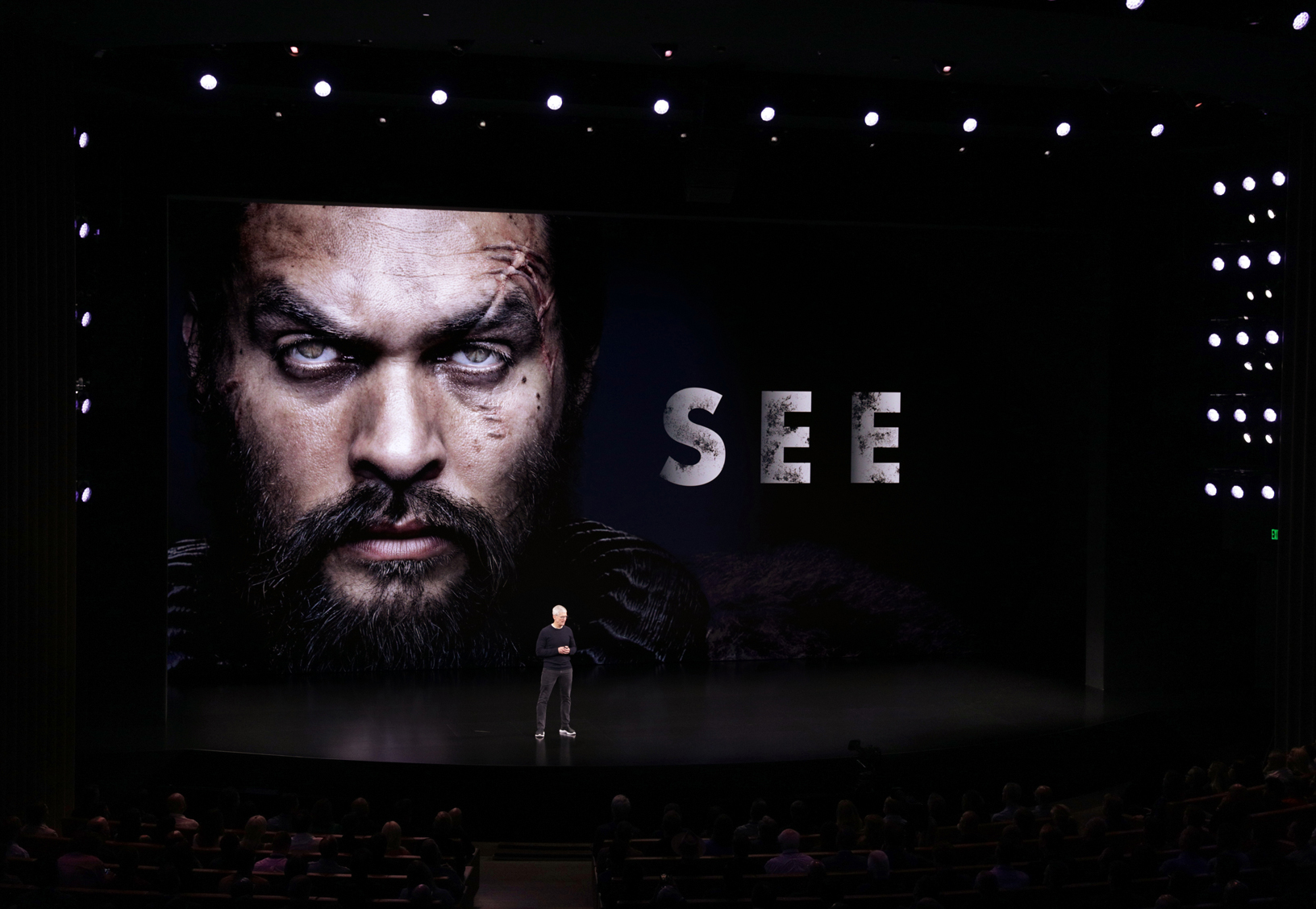 Tim Cook stands on a dark stage with a large image of actor Jason Momoa and the word See on a screen behind Cook.