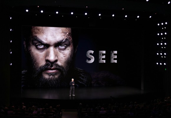 Tim Cook stands on a dark stage with a large image of actor Jason Momoa and the word See on a screen behind Cook.