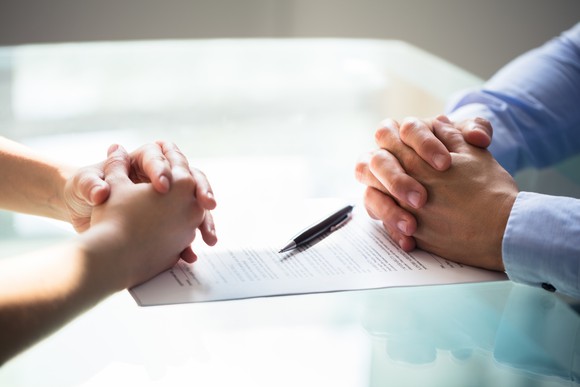 Two businesspeople folding their hands on a glass table, separated by a pen and a document.