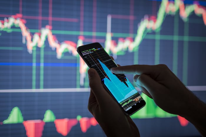 A hand touching a smartphone screen showing a stock chart, with an even bigger stock chart on the wall behind it. 