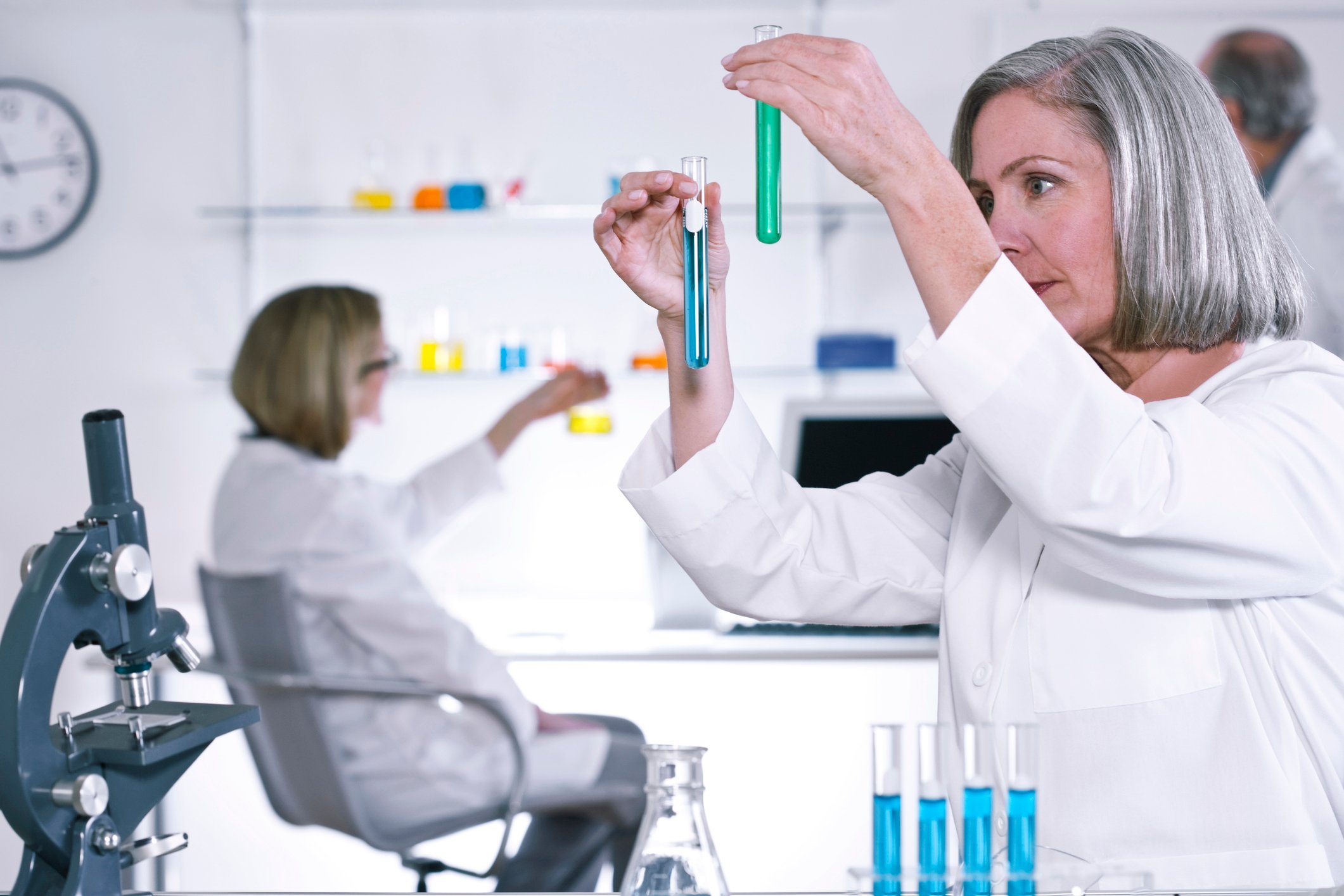 Female scientist holding two test tubes with a microscope and test tube rack nearby and other scientists in the background