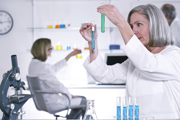 Female scientist holding two test tubes with a microscope and test tube rack nearby and other scientists in the background