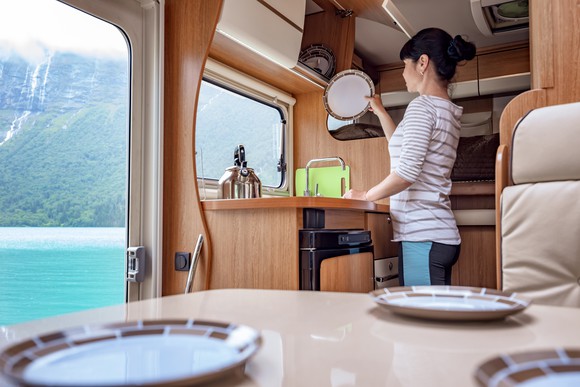 A woman washes dishes in her RV kitchen in view of a mountain lake.