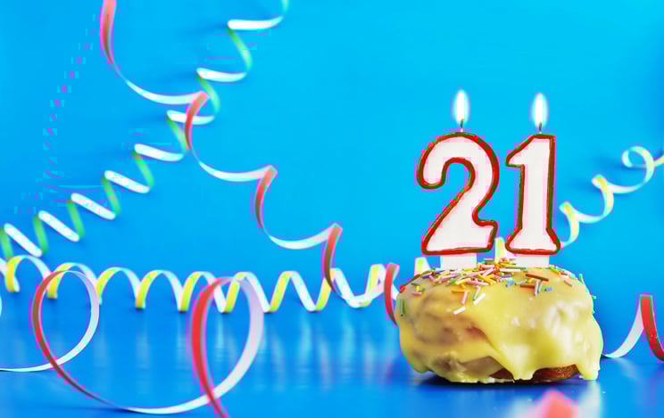 Two lit candles -- a 2 and a 1 -- in a cupcake surrounded by colorful curly paper streamers on a blue background.