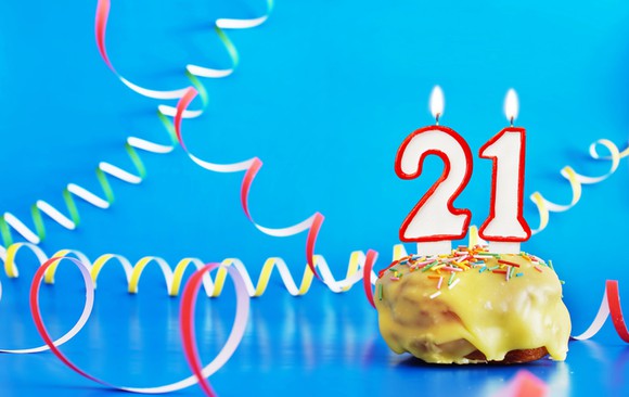 Two lit candles -- a 2 and a 1 -- in a cupcake surrounded by colorful curly paper streamers on a blue background.