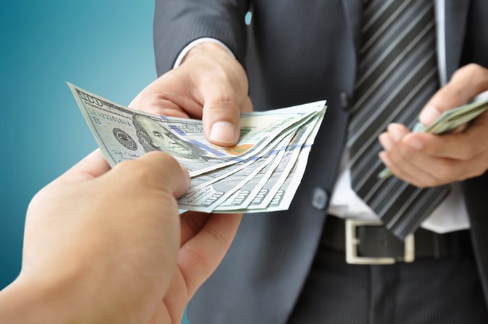 A businessman in a suit holding a stack of cash in his left hand while handing over a few one hundred dollar bills in his right hand to another person's hand.