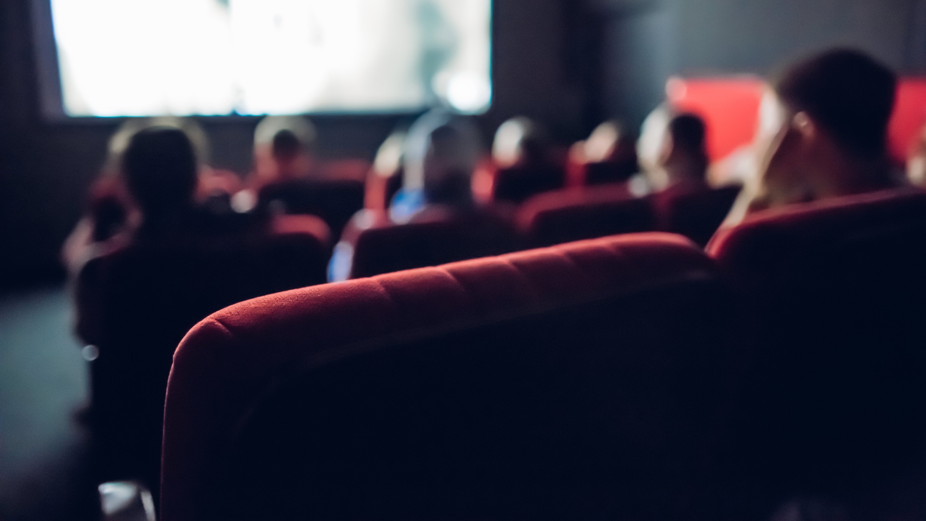 Close-up of the back of a seat in a movie theater with the screen visible out of focus beyond it. 