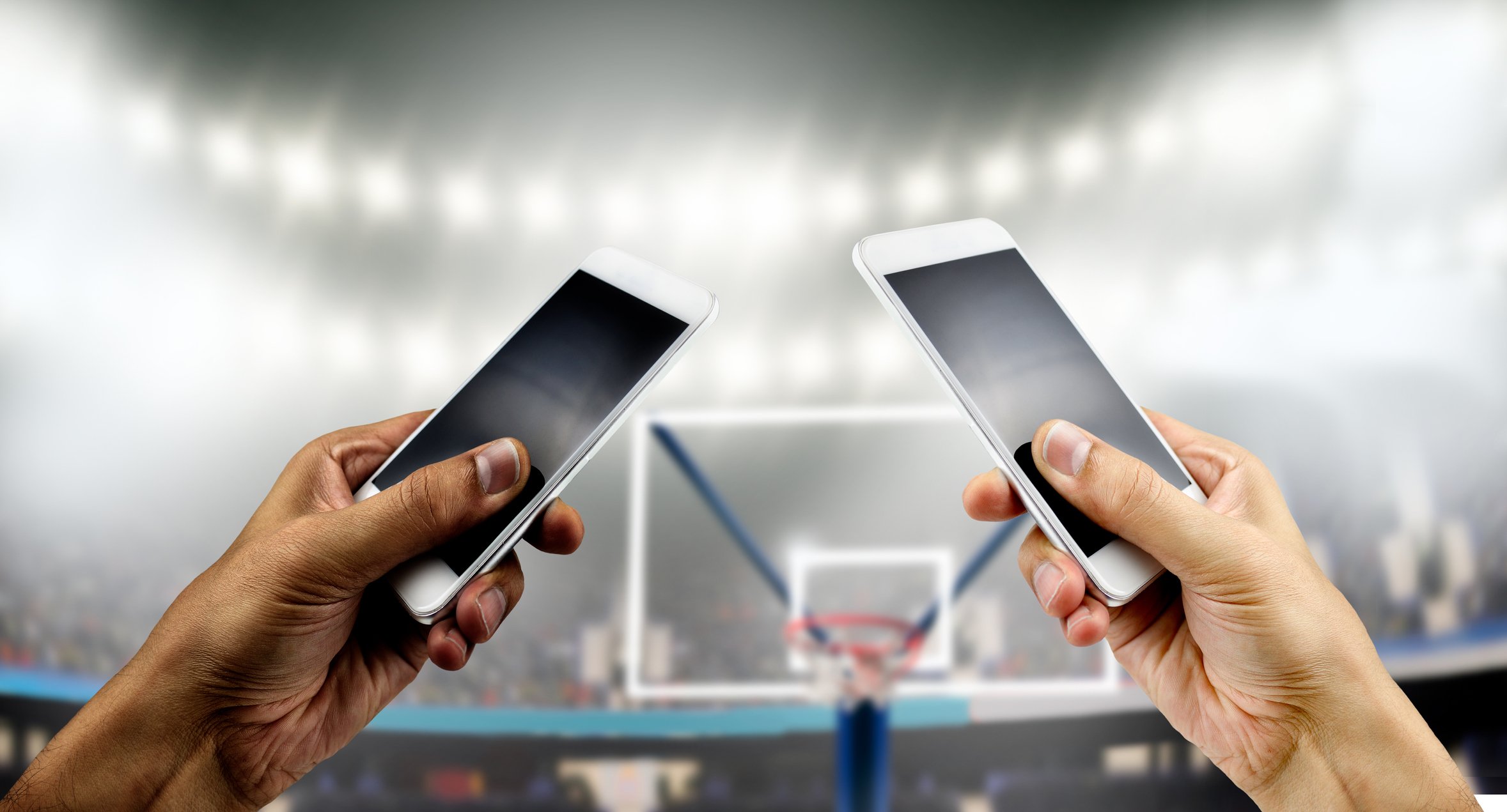 Two different hands, each holding a smartphone, in front of a basketball hoop. 
