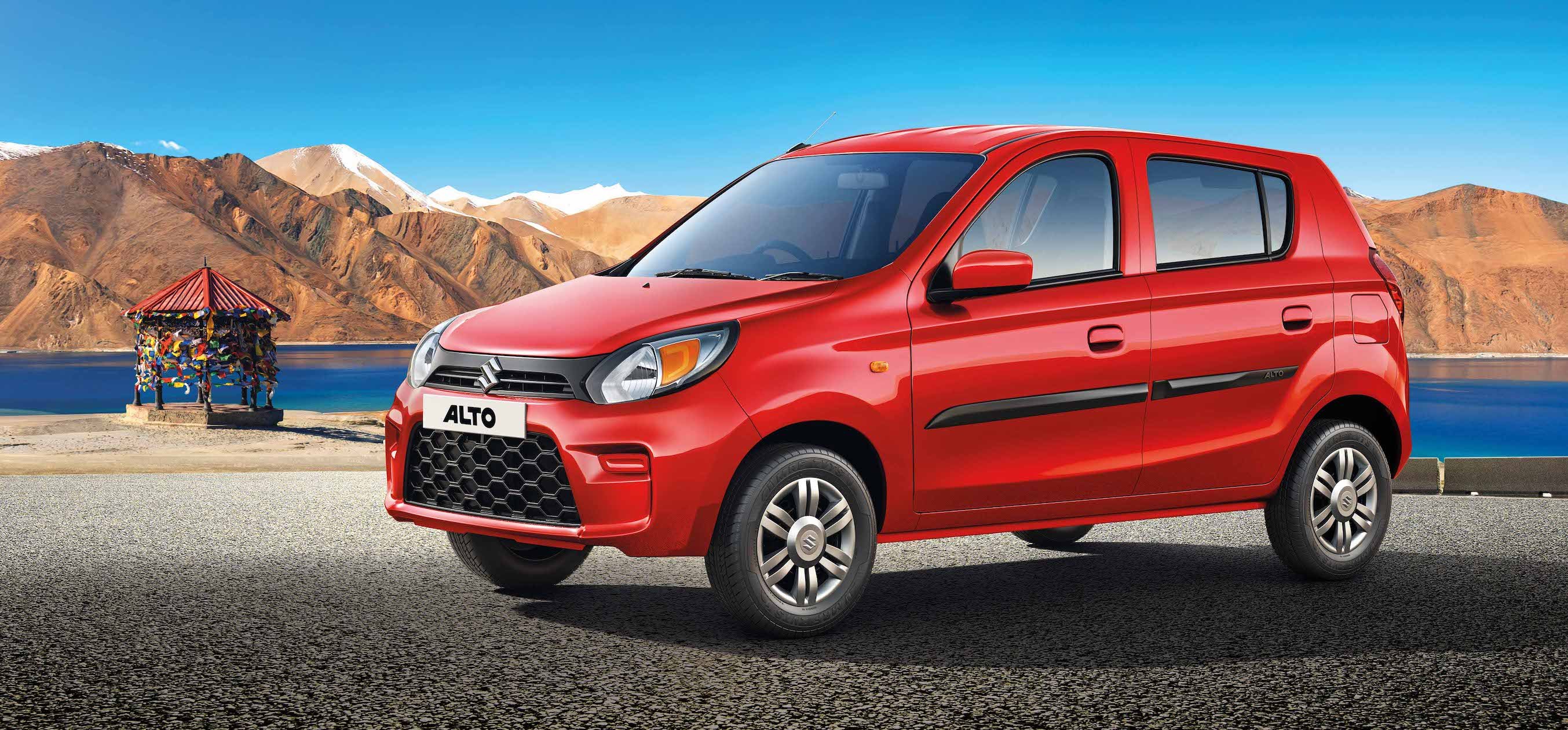 A red Suzuki Alto parked in front of a lake with mountains in the background
