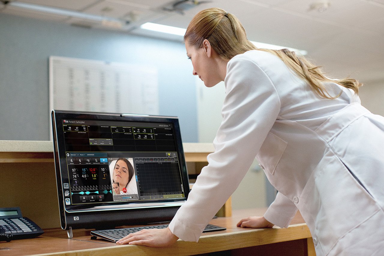 A woman in white doctor's labcoat looking at a computer screen displaying vitals and a woman sleeping.