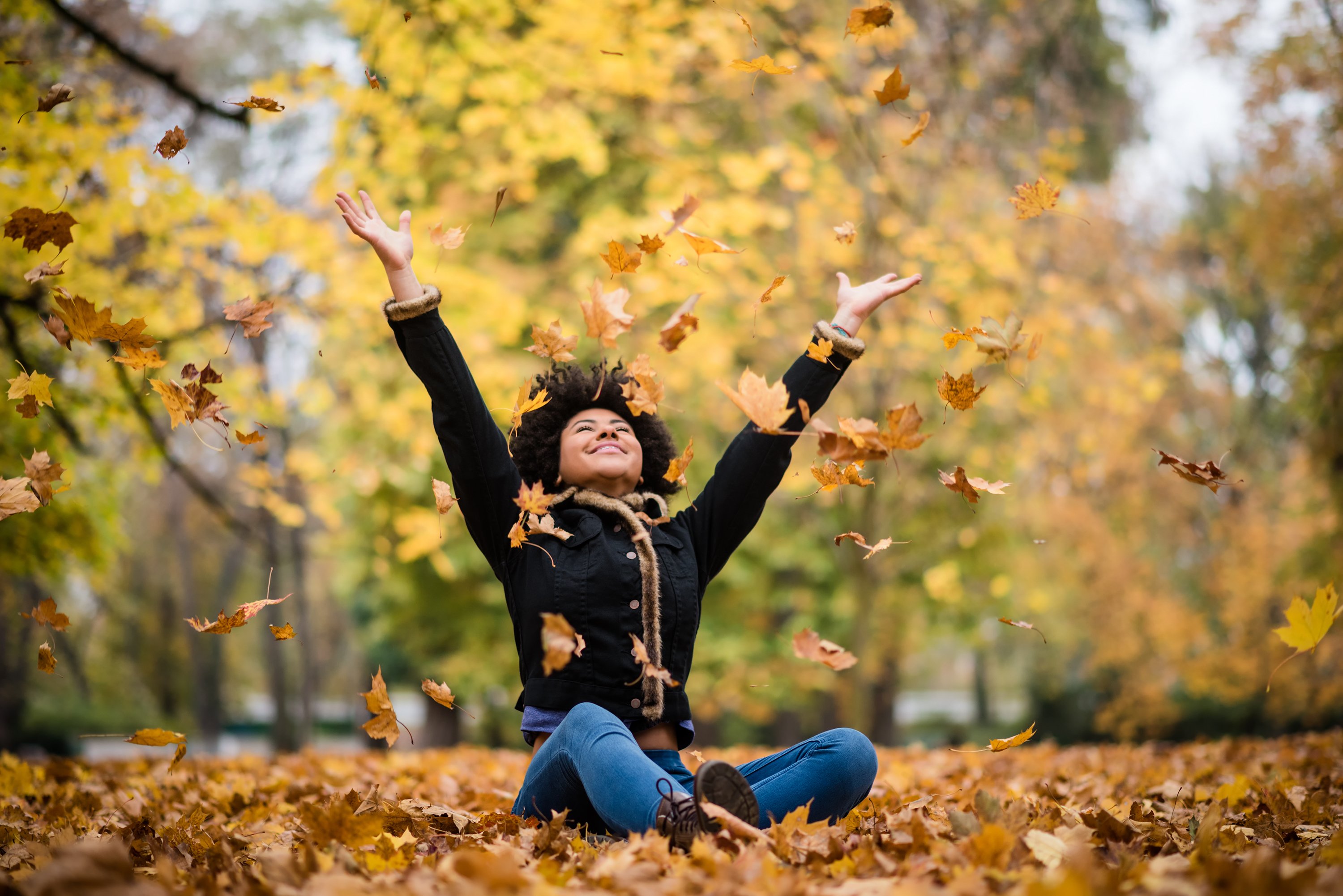 A woman sits on ground covered in leaves and throws leaves up in the air in joy. 