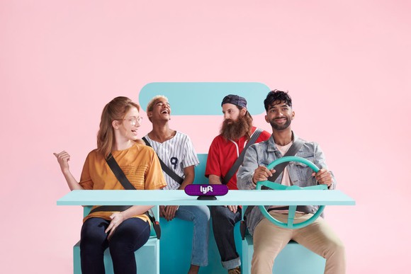 Four young riders in a restaurant booth made to look like an imaginary Lyft car, with a steering wheel and seatbelts