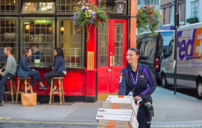 A female FedEx worker pushing a box of FedEx boxes with a FedEx truck in the background