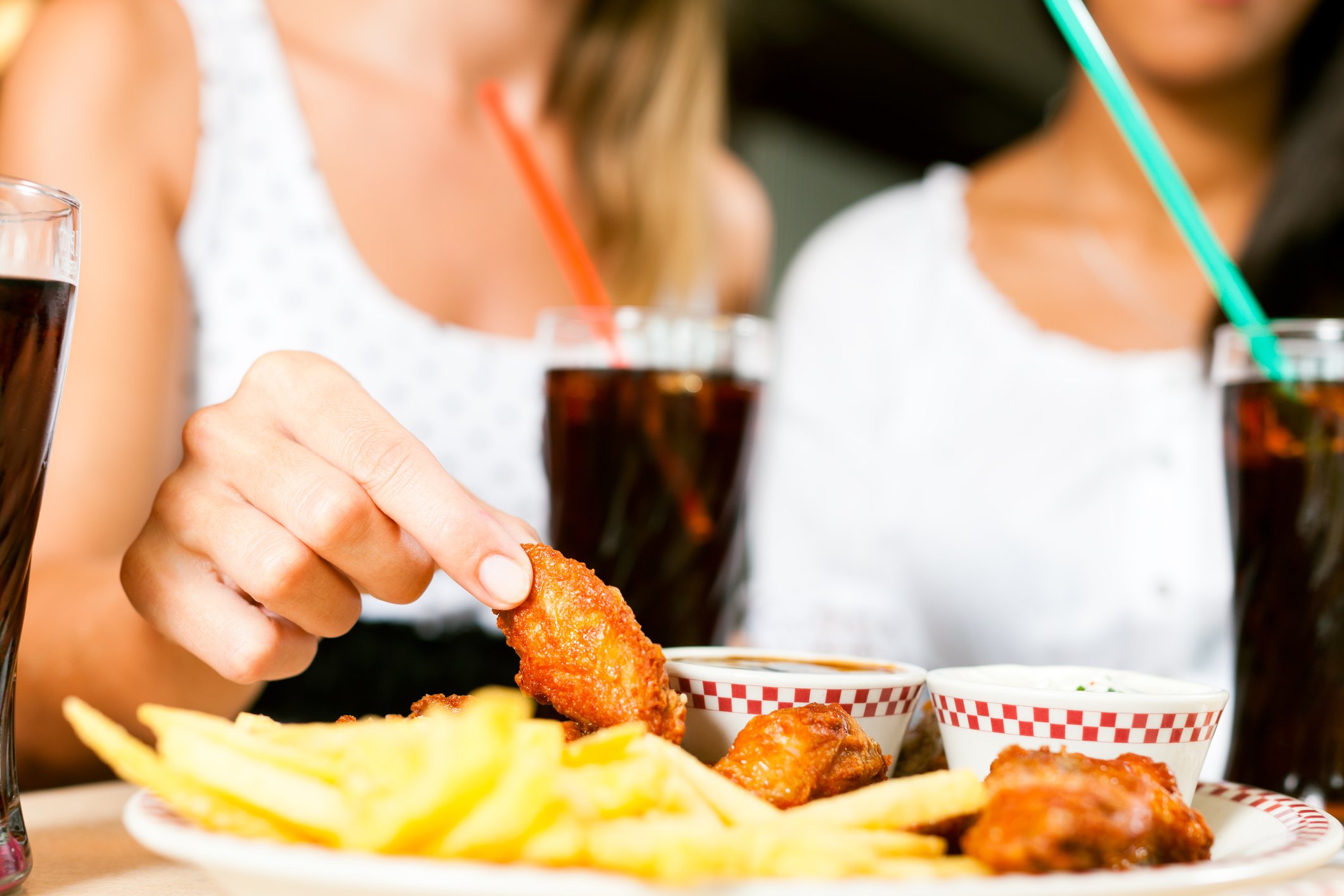 Two women eating chicken wings with sodas.