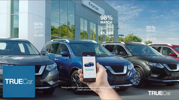 Hand holding up a smartphone displaying the TrueCar app with four cars in the background