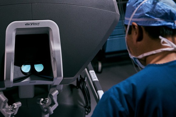 A surgeon wearing a mask sitting next to a da Vinci machine and viewing lens for the procedure.
