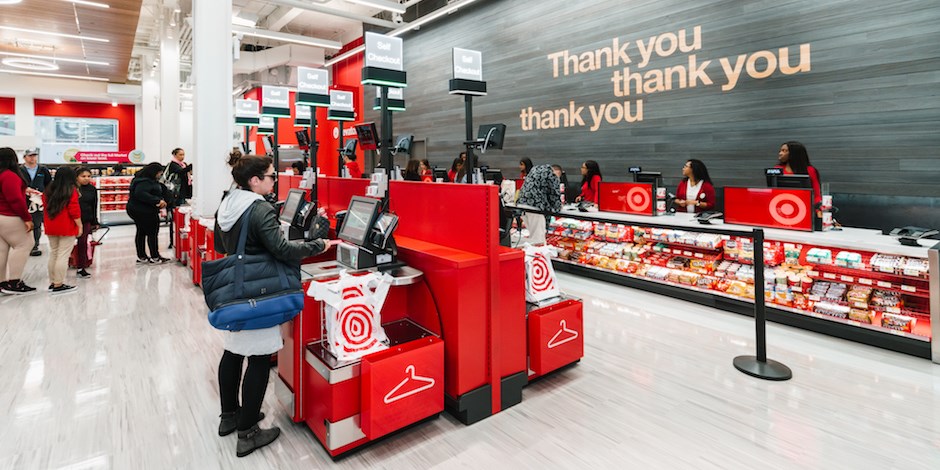 The interior of a Target store. Digital checkout kiosks with people making purchases are in the foreground with a customer service counter in the background.