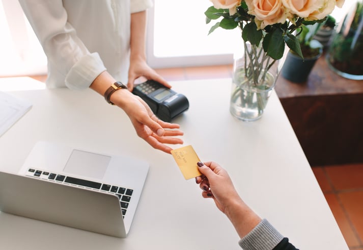 A woman's hand handing a credit card to a woman behind a desk that has a payment processing device and laptop on it.